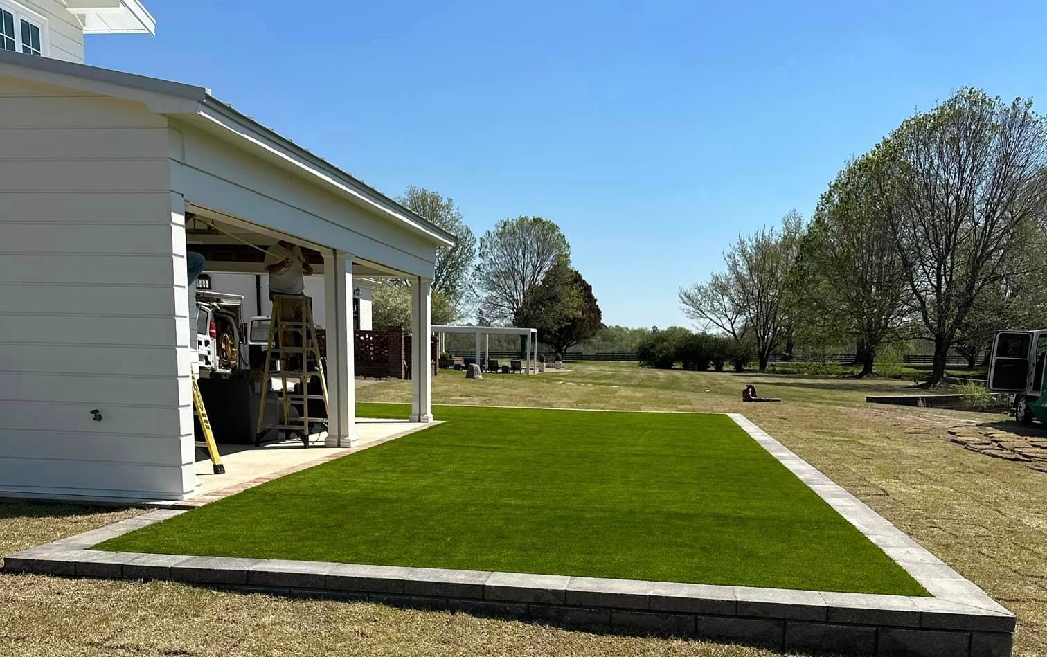 A rectangular patch of green artificial turf is bordered by a low retaining wall next to a house under a clear sky.