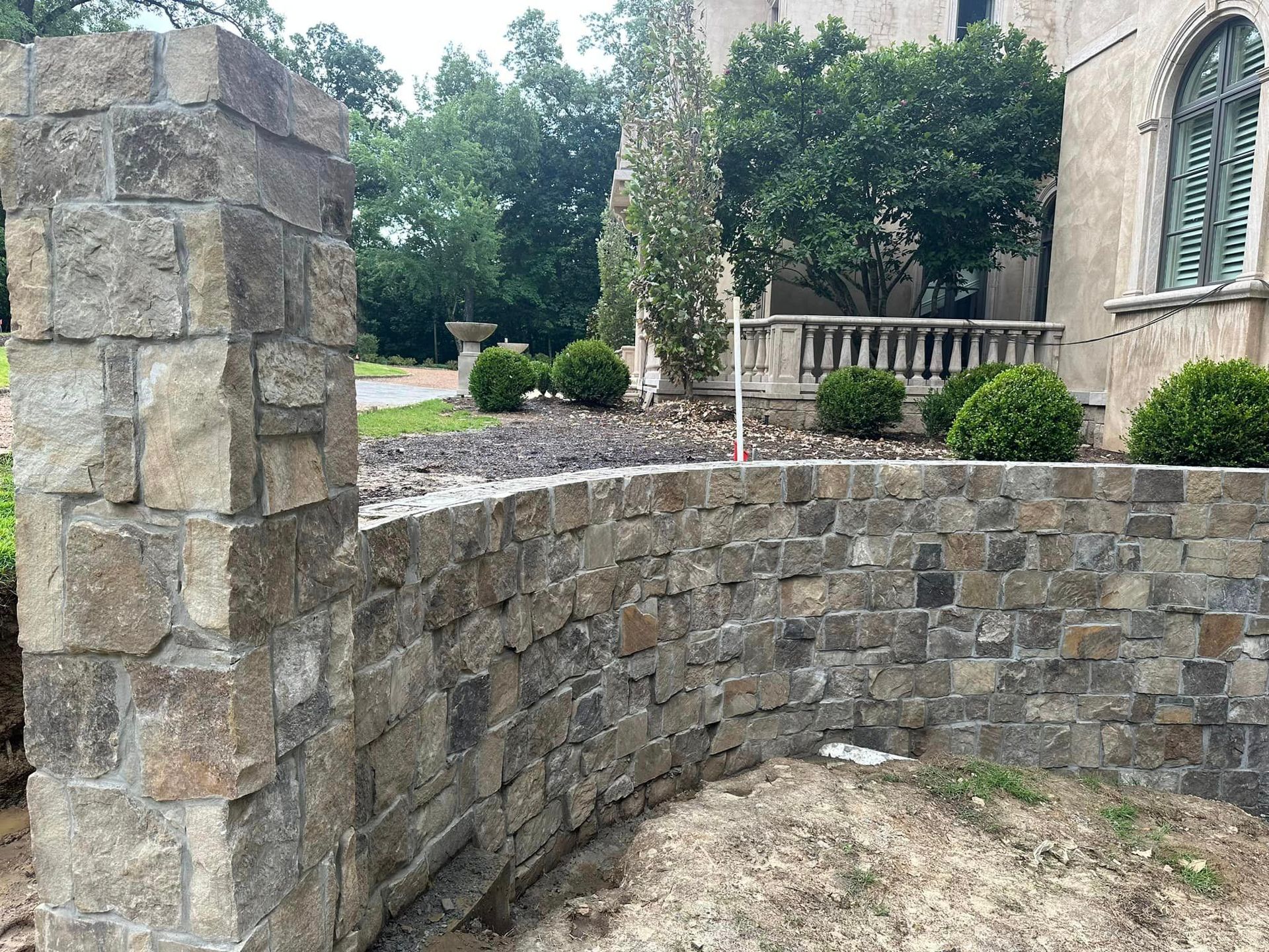 Stone wall and pillar construction with a building and greenery in the background.
