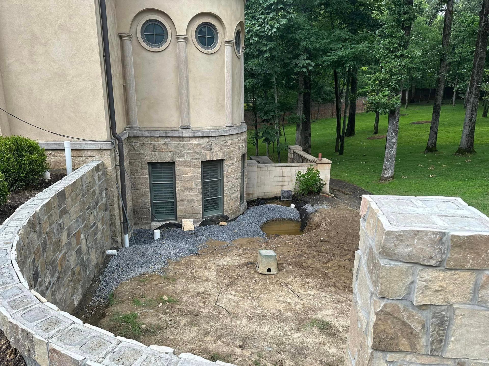 Exterior of a building with stone walls and a grassy area under construction. Trees in the background.