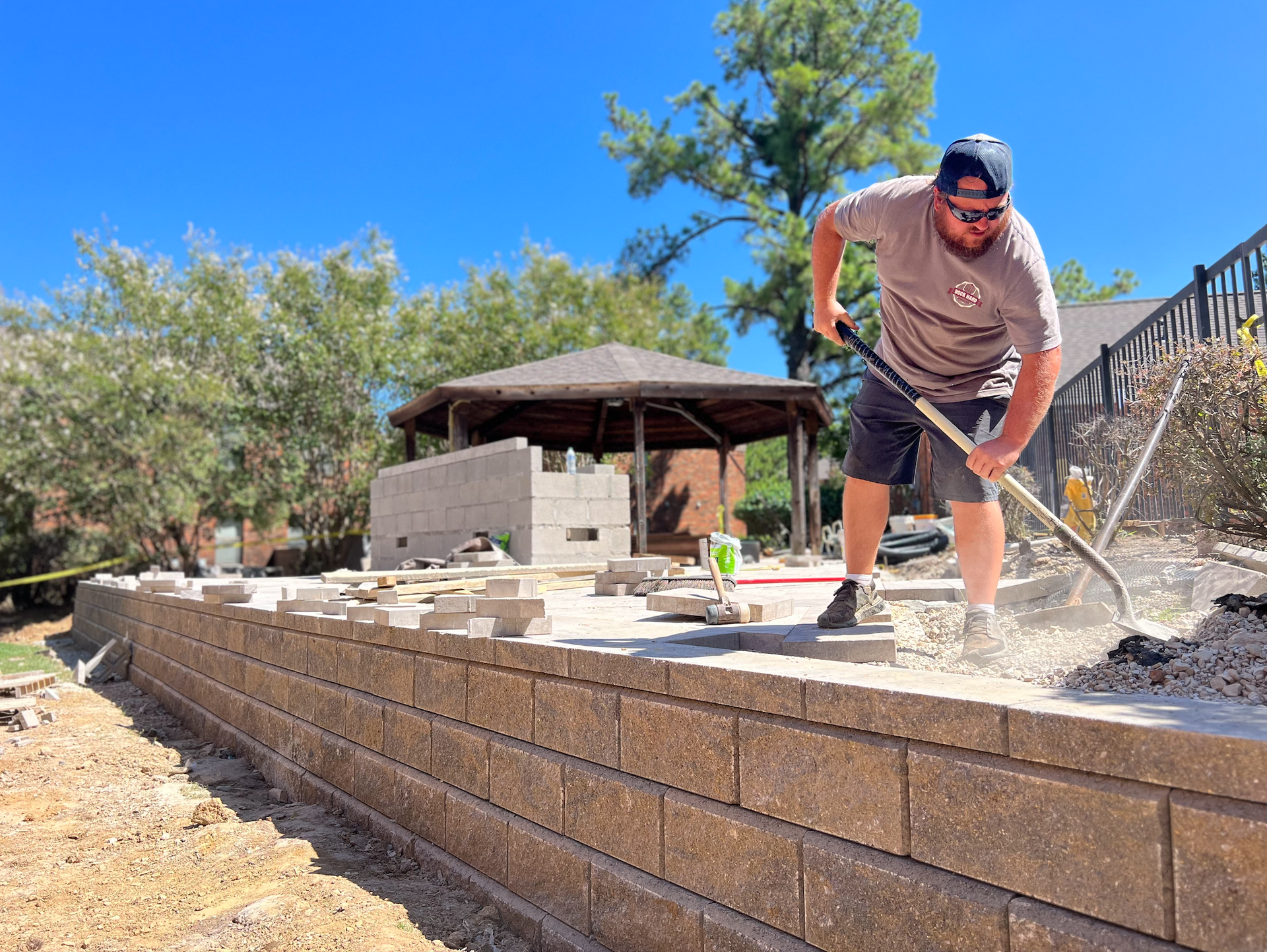 A man raking gravel on a patio under construction, with a retaining wall and gazebo in the background.