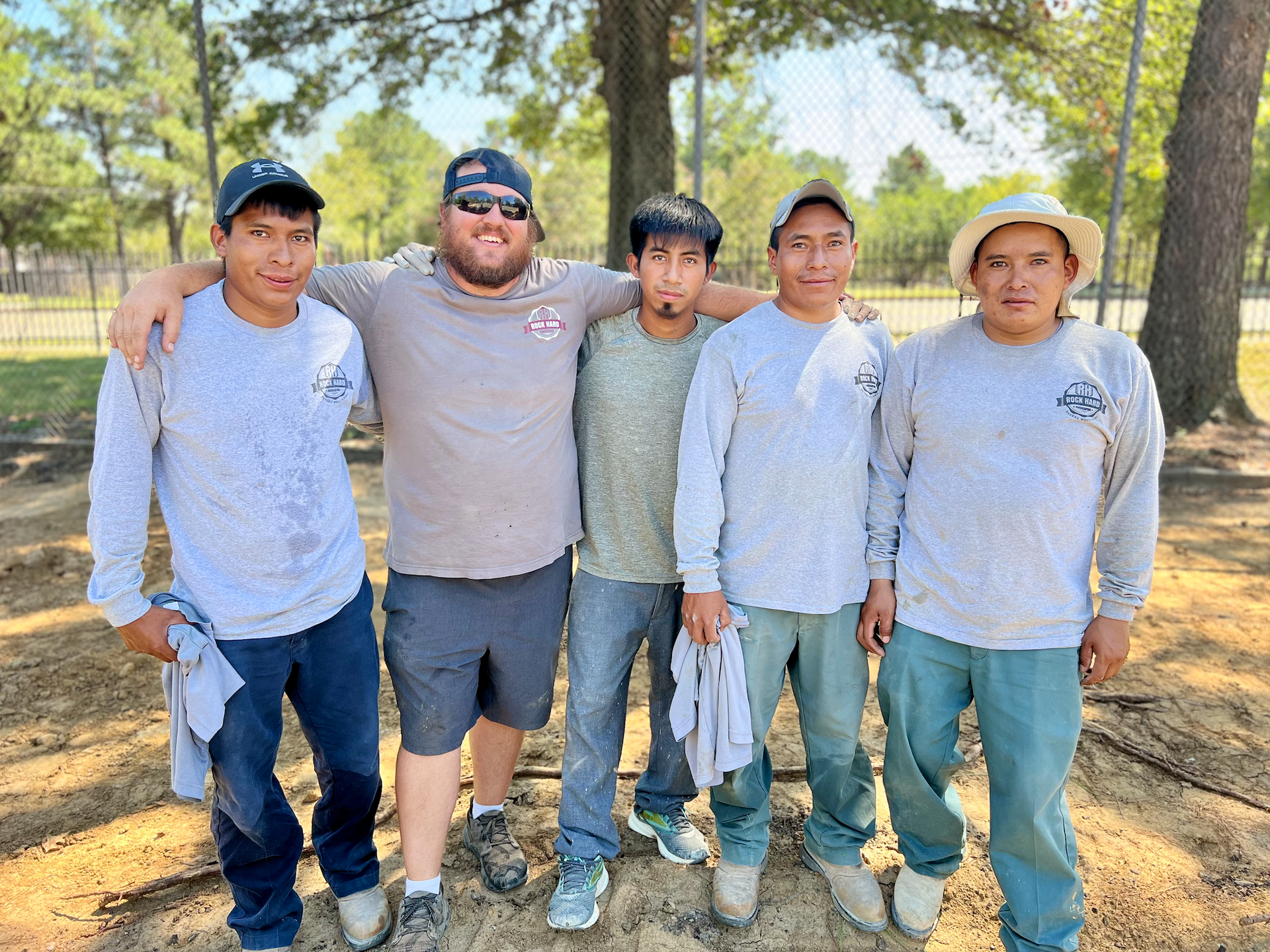 Five people standing together outdoors; four in matching gray shirts, one with an arm around two of them.