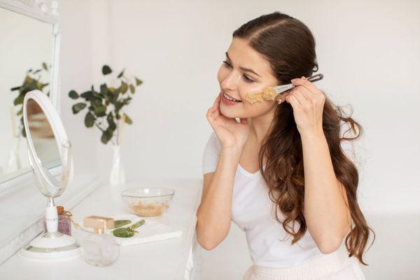 Mujer aplicándose una mascarilla facial con brocha frente al espejo.