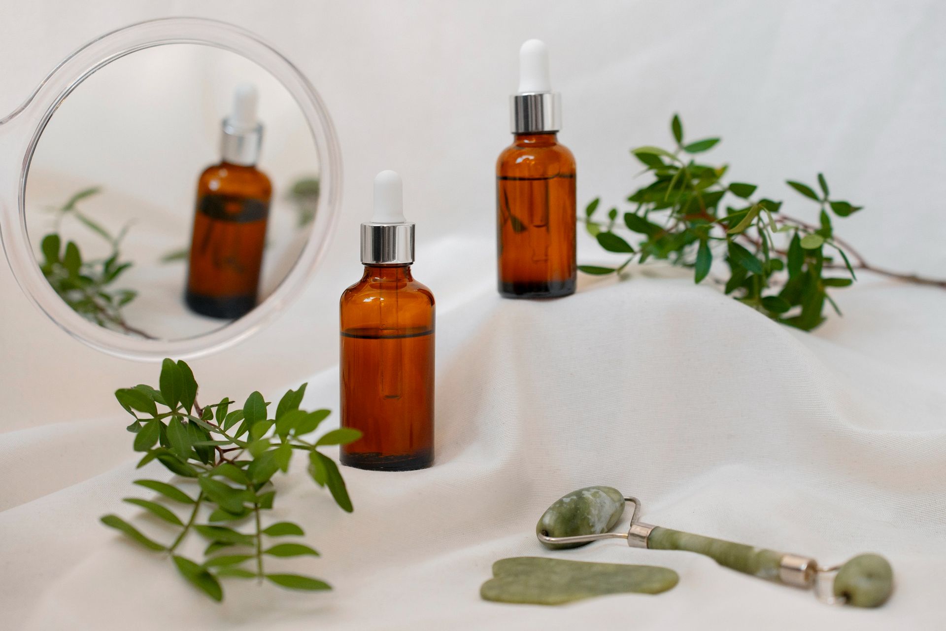 Dos botellas de vidrio ámbar, un rodillo de jade y una gua sha sobre tela blanca con vegetación.