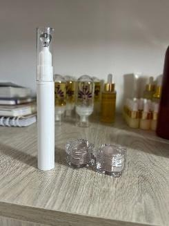 Tubo cosmético blanco con aplicador de bolita y dos frascos pequeños sobre una superficie de madera. Otras botellas al fondo.