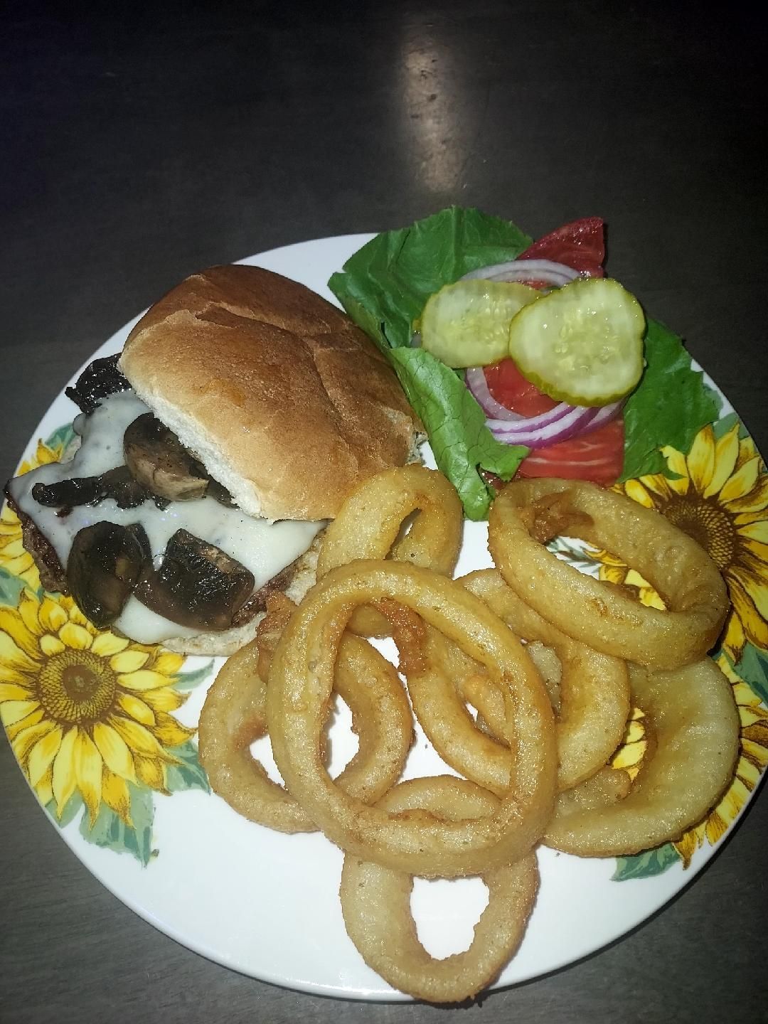 Burger with onion rings, pickles, lettuce, and tomato on a sunflower-decorated plate.
