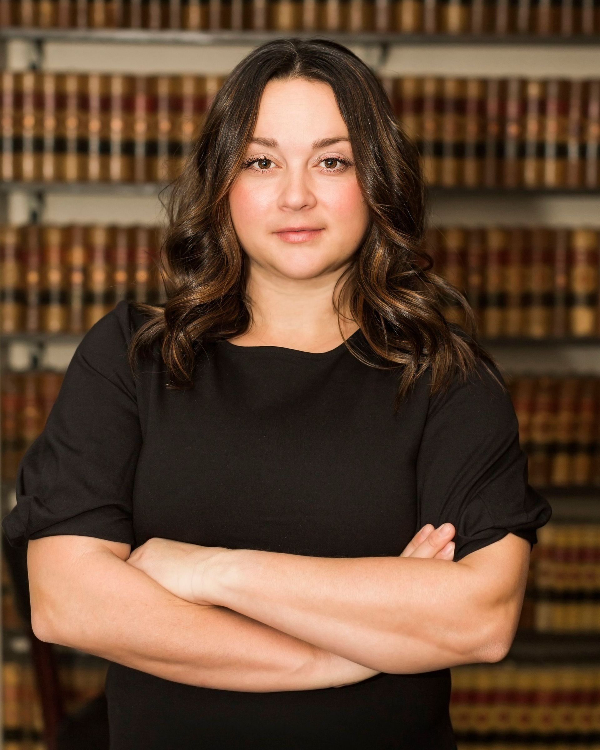 A woman in a black dress is standing with her arms crossed in front of a bookshelf.