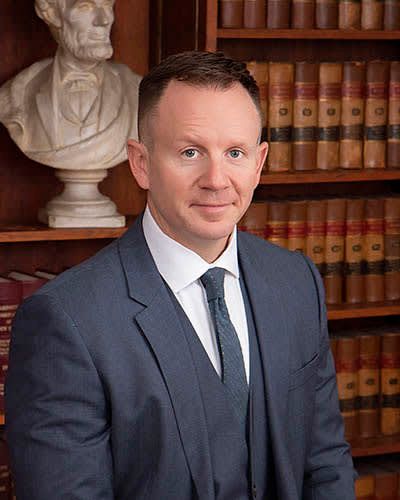 A man in a suit and tie is standing in front of a bookshelf.