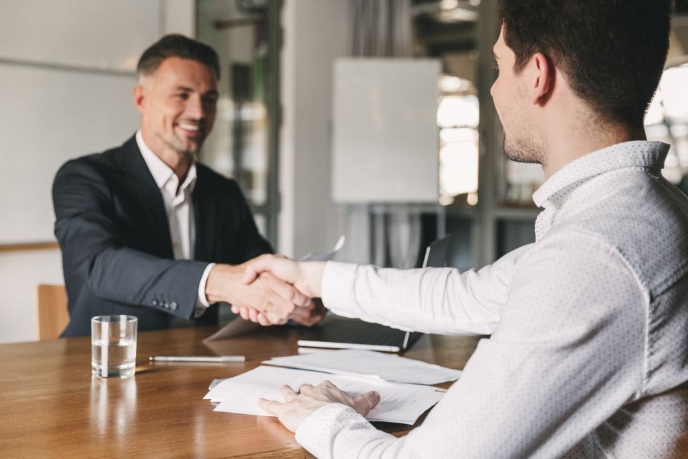 Two Men Are Shaking Hands While Sitting at a Table — All About Conveyancing Taree In Taree, NSW