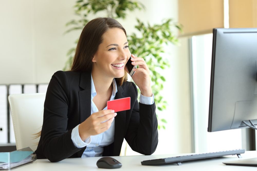 A Woman is Sitting at a Desk Holding a Credit Card and Talking on a Cell Phone — All About Conveyancing Taree In Old Bar, NSW