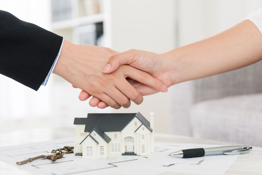 A Man and a Woman Are Shaking Hands in Front of a Model House — All About Conveyancing Taree In Taree, NSW