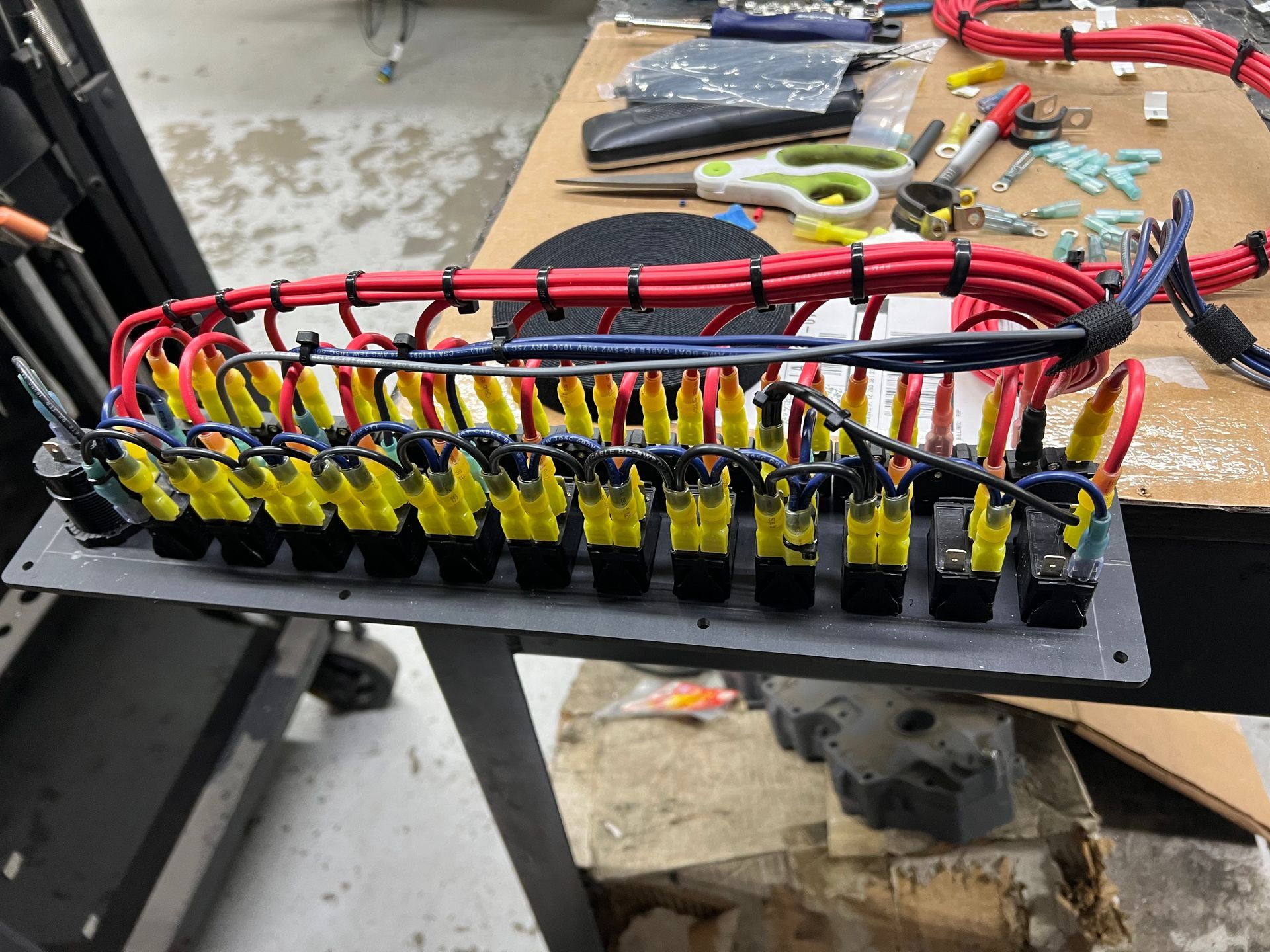 Wiring panel with many yellow-capped fuses and red and black wires, set on a workbench.