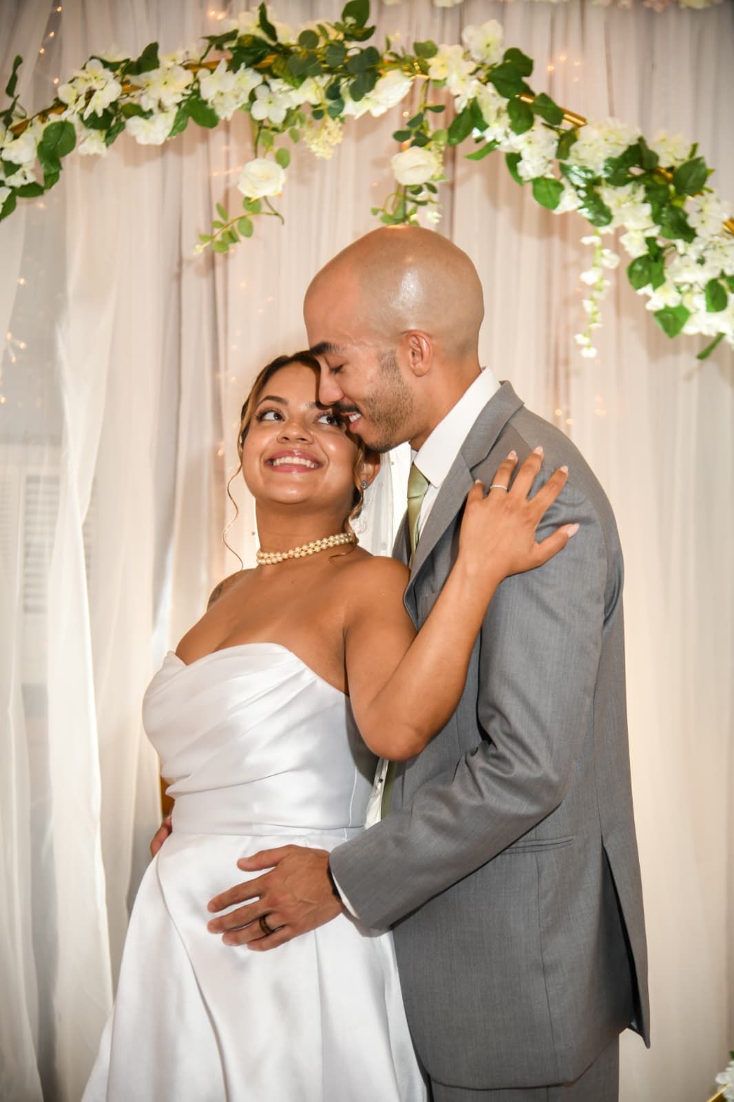 A smiling bride in a white wedding gown and a groom in a grey suit standing together under a floral arch.