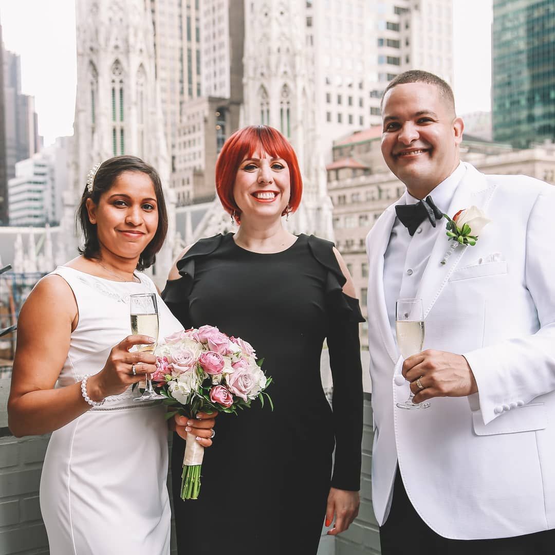Three people pose with champagne glasses and a bouquet on a rooftop, with a city building visible in the background.