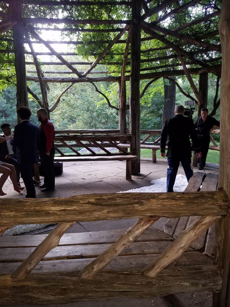 People in formal attire gather under a rustic, open-air wooden gazebo in a lush park setting.