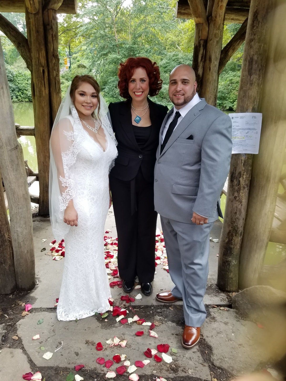 A bride, groom, and officiant stand together under a wooden pavilion, surrounded by rose petals scattered on the ground.