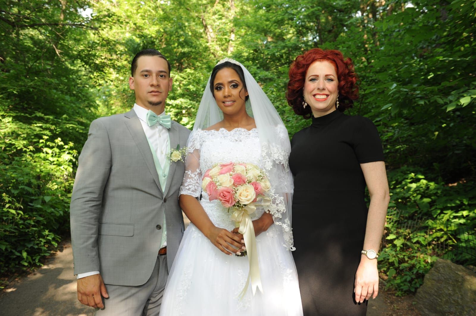 A bride in a white wedding gown holding a bouquet, a groom in a grey suit, and a woman in a black dress stand in a forest.