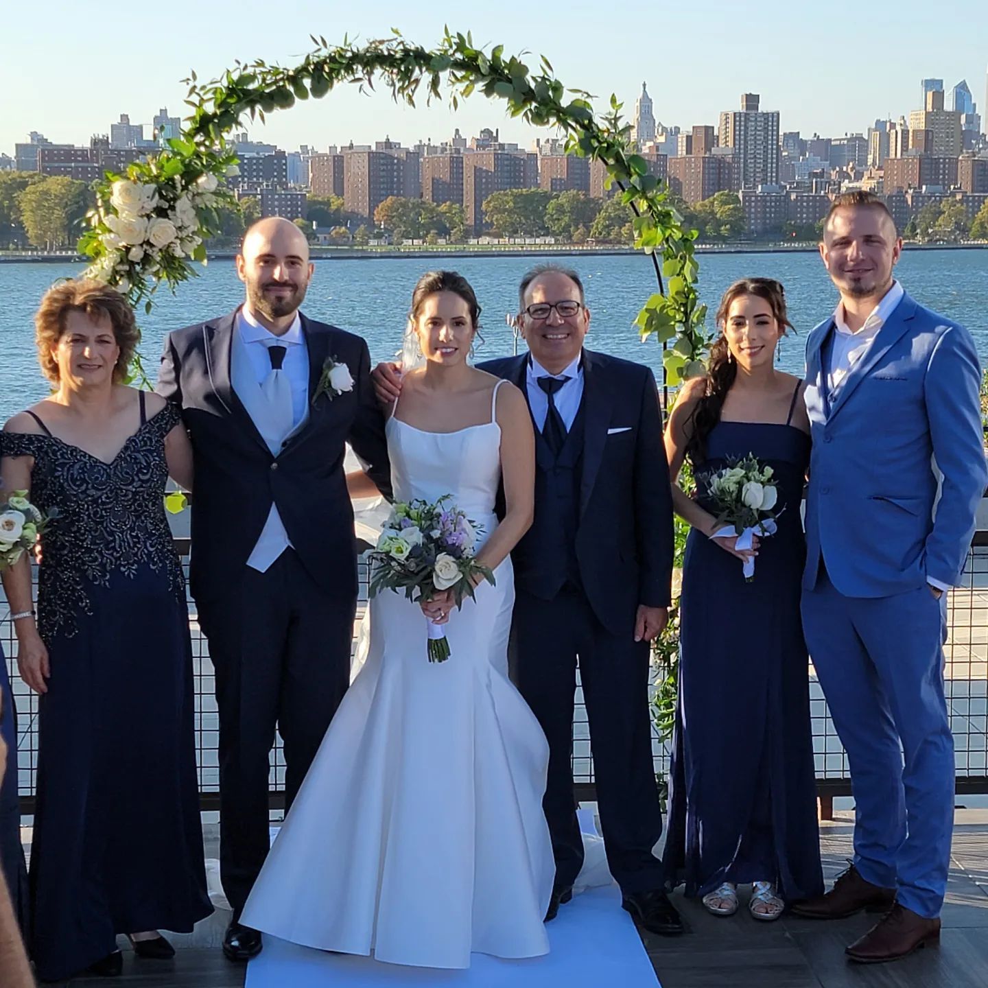Wedding group portrait by water: Bride and groom stand under floral arch with family, cityscape background.