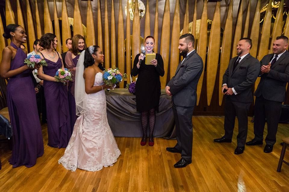 Wedding ceremony. Bride and groom face officiant; bridal party and groomsmen watch. Wooden backdrop.