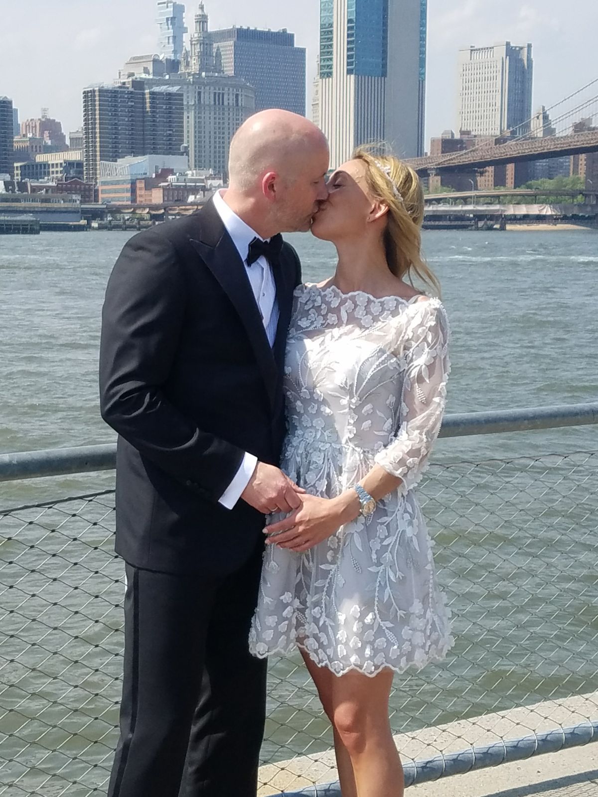 A couple in wedding attire kisses near a waterfront railing with the New York City skyline in the background.