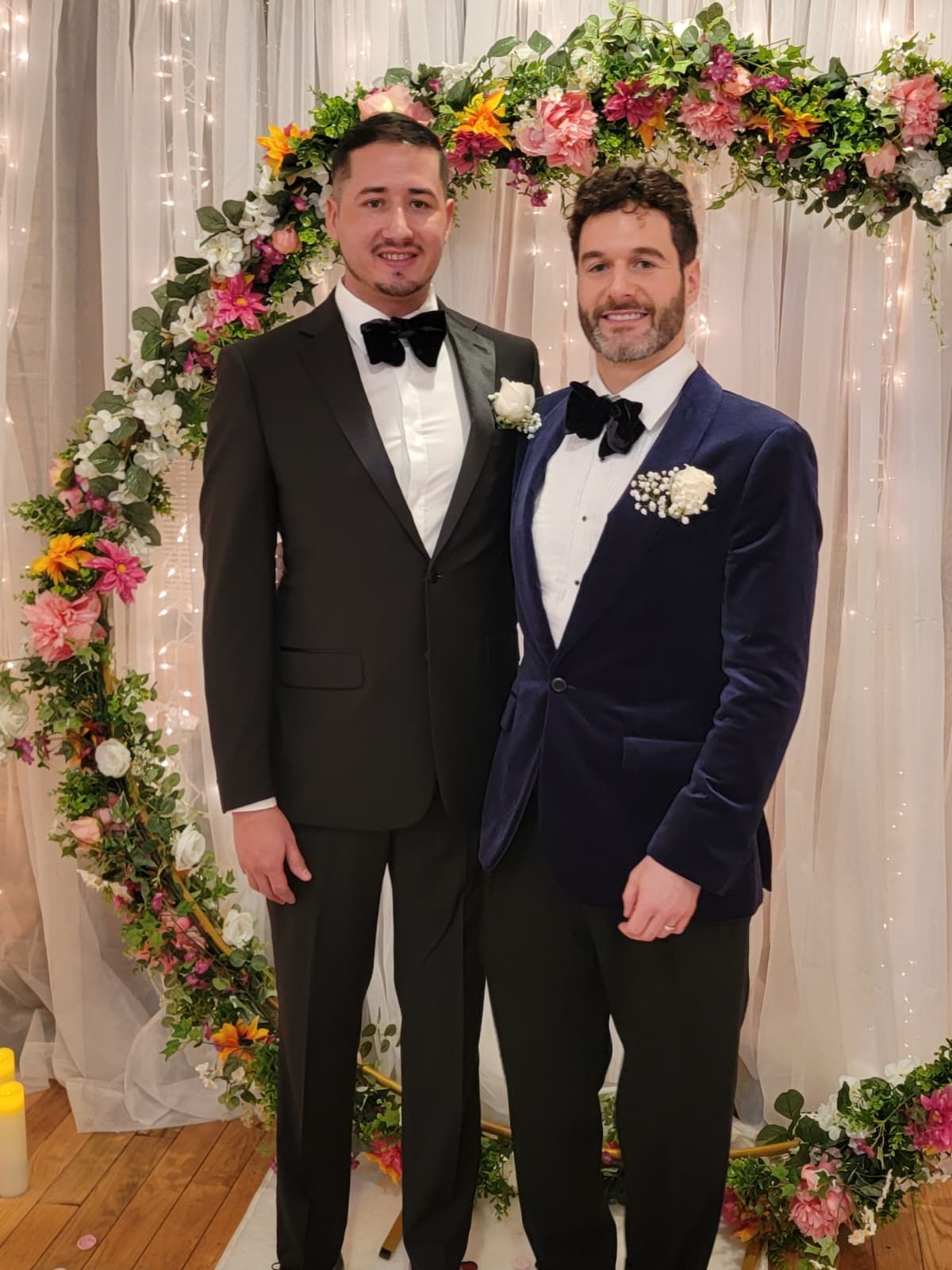 Two individuals in formal tuxedos and bow ties smiling together in front of a floral wedding arch and draped backdrop.