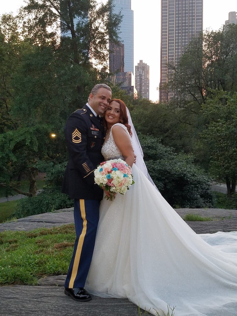 A person in a military dress uniform and a person in a white wedding gown holding a bouquet in a park with city buildings.