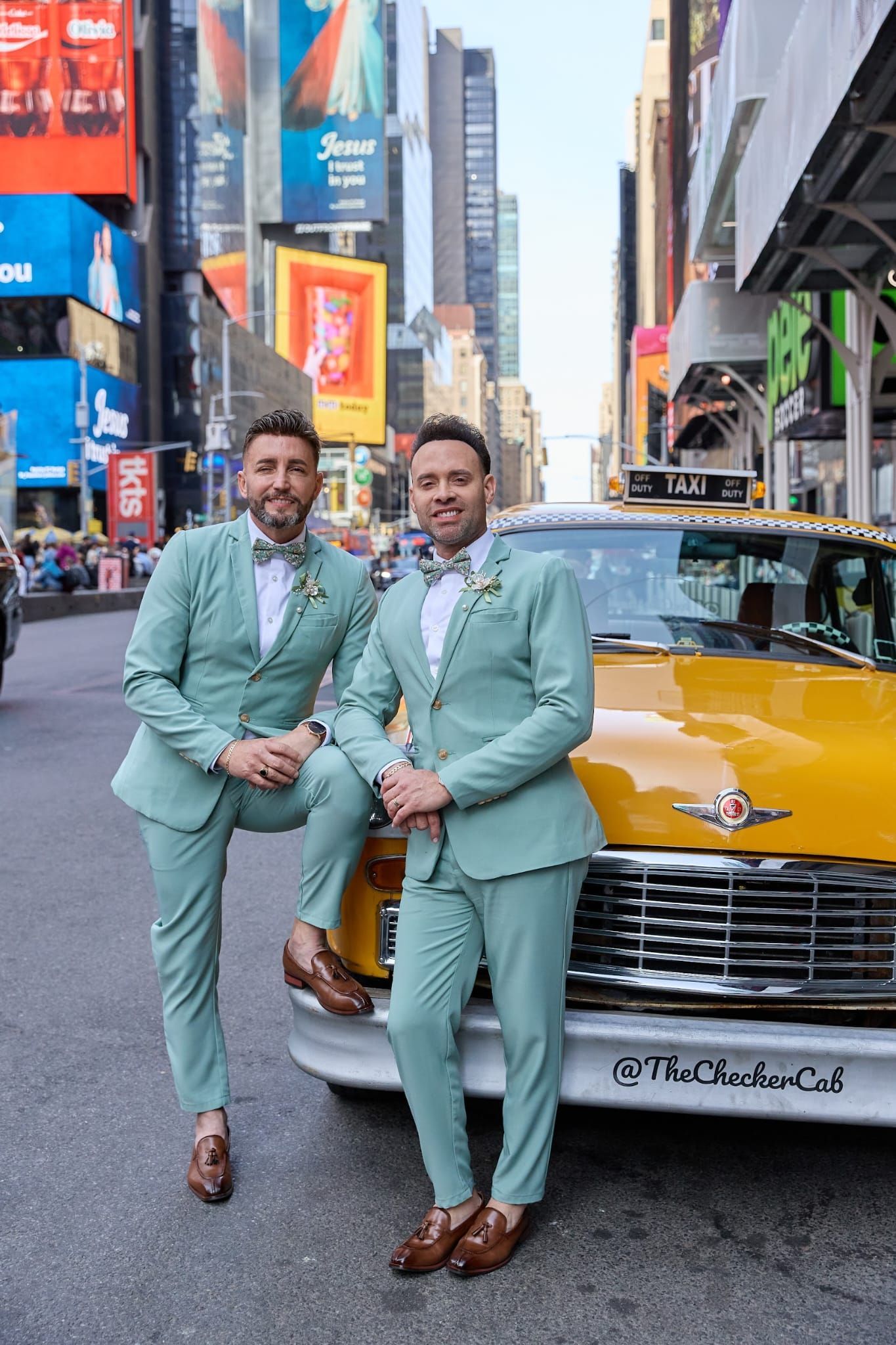 Two people in matching sage green suits pose next to a yellow taxi in Times Square.