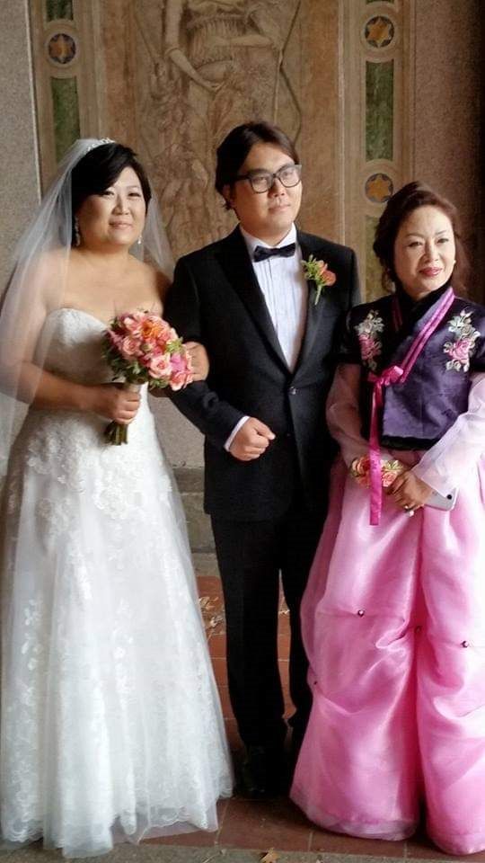 A bride in a white wedding gown holding a bouquet, a groom in a suit, and a woman in a purple and pink hanbok.