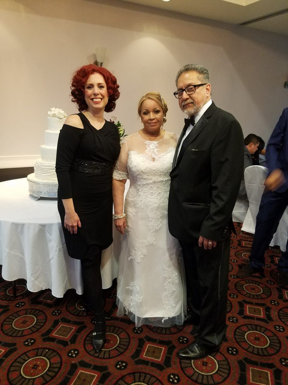 Three people pose for a photo at a formal event near a tiered wedding cake.