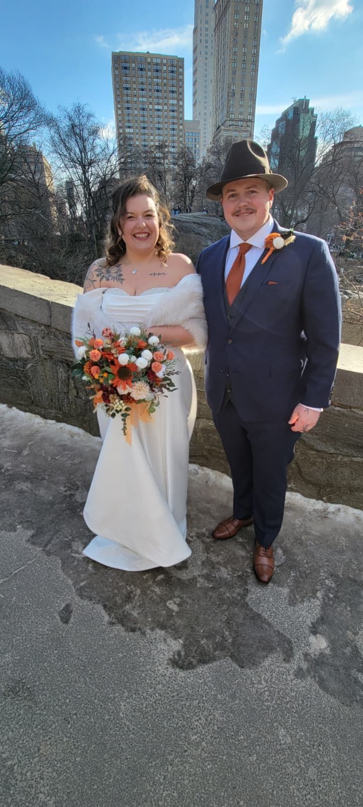 Bride signs document on bench with smiling man in suit and woman in colorful dress.