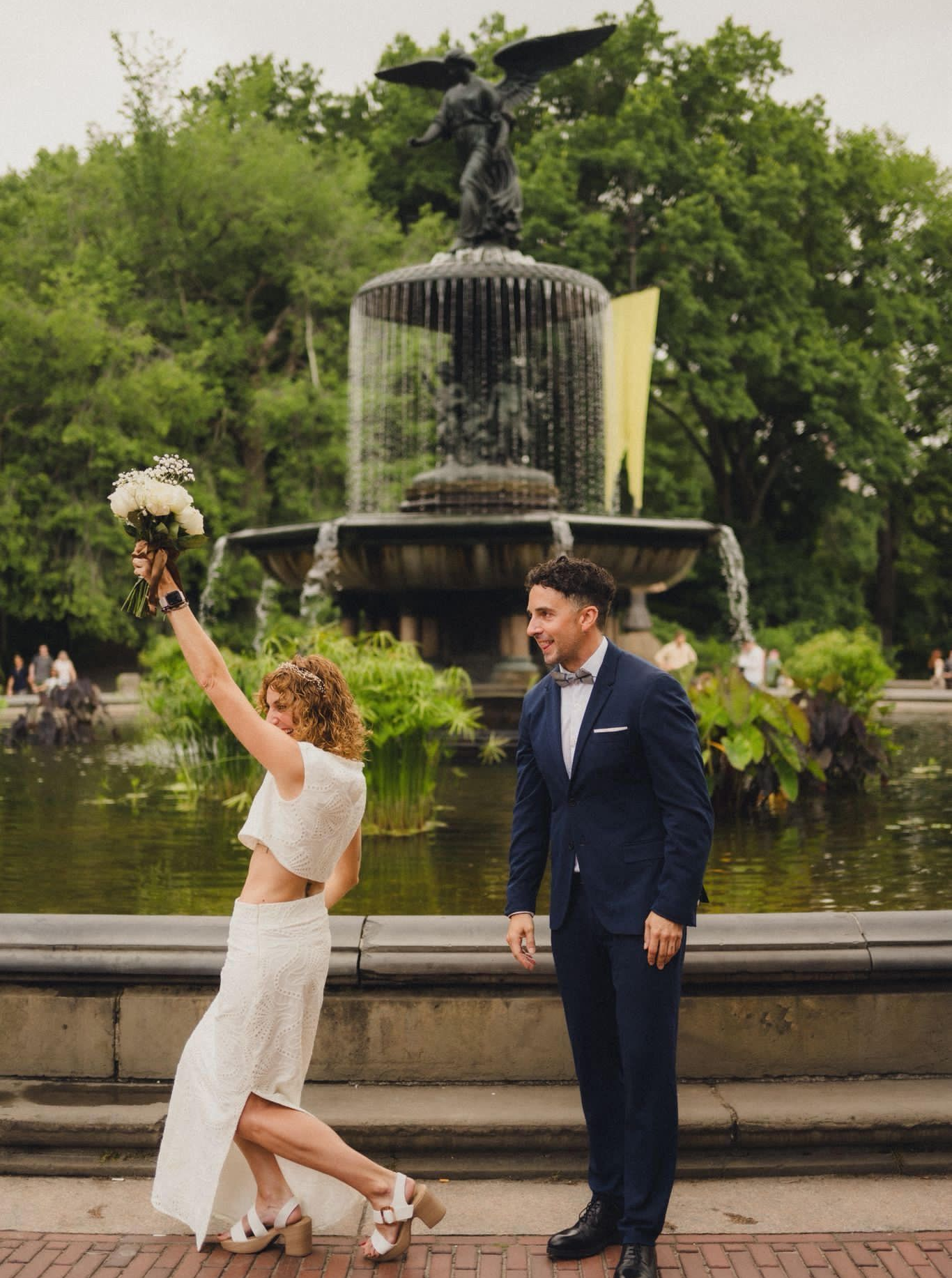 A person in a white two-piece wedding outfit holds a bouquet aloft while standing before the Bethesda Fountain in NYC.