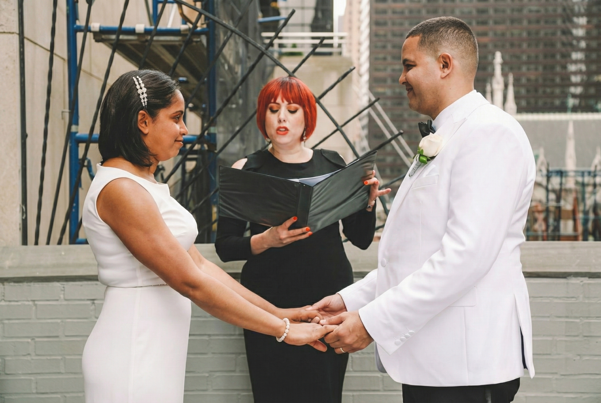 A couple in white formal attire holds hands while an officiant in black reads from a folder during an outdoor ceremony.