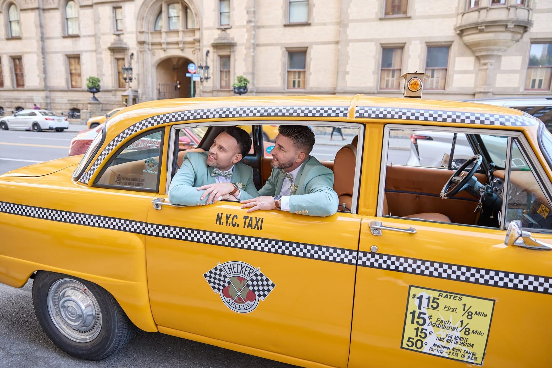 Two people in light green suits lean out the window of a yellow checker taxi in front of a historic building.