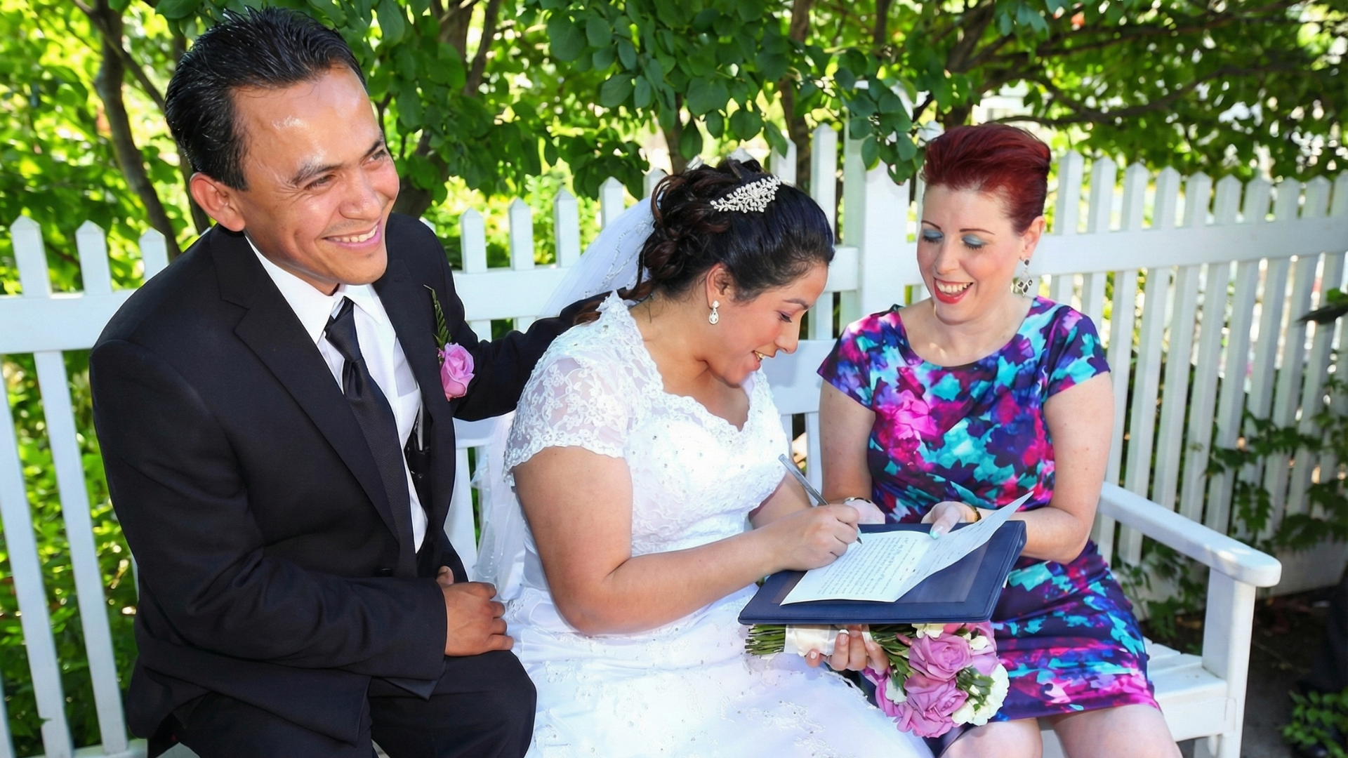 A bride in a white wedding dress signs a document, seated on a bench beside her partner and an officiant outdoors.
