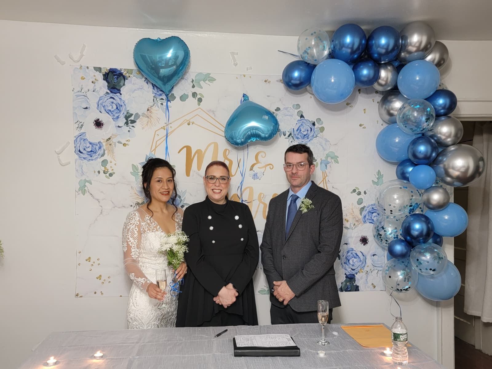 Wedding party poses on a green lawn with a white pergola in the background. Women in gowns, men in suits.