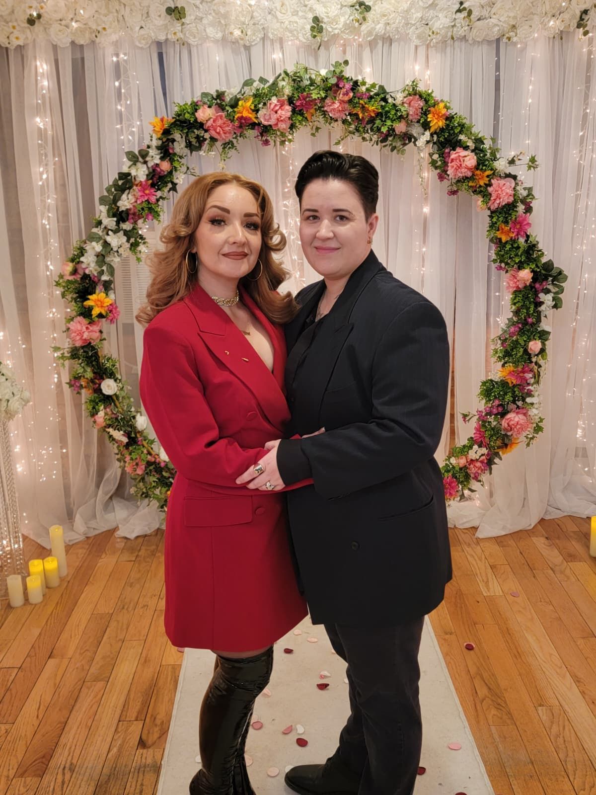Wedding ceremony: Groom placing ring on bride's finger, officiant smiling, indoor setting with floral backdrop.
