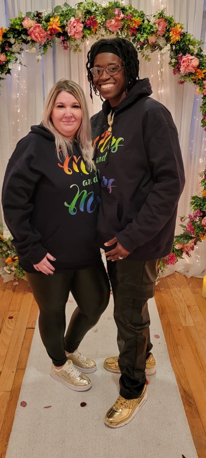 Two people smile, wearing matching black hoodies with colorful text and gold sneakers, standing before a floral arch.