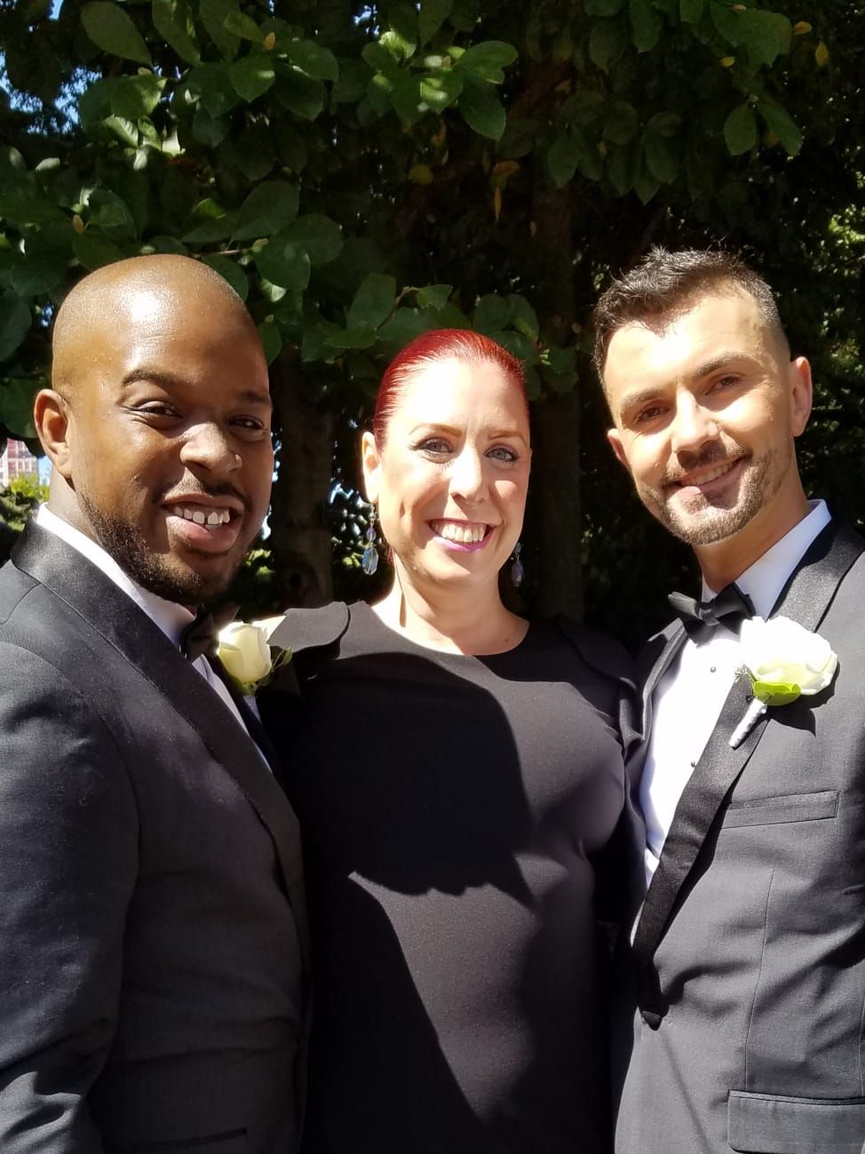 Three people in formal attire stand smiling side-by-side in front of green foliage on a sunny day.