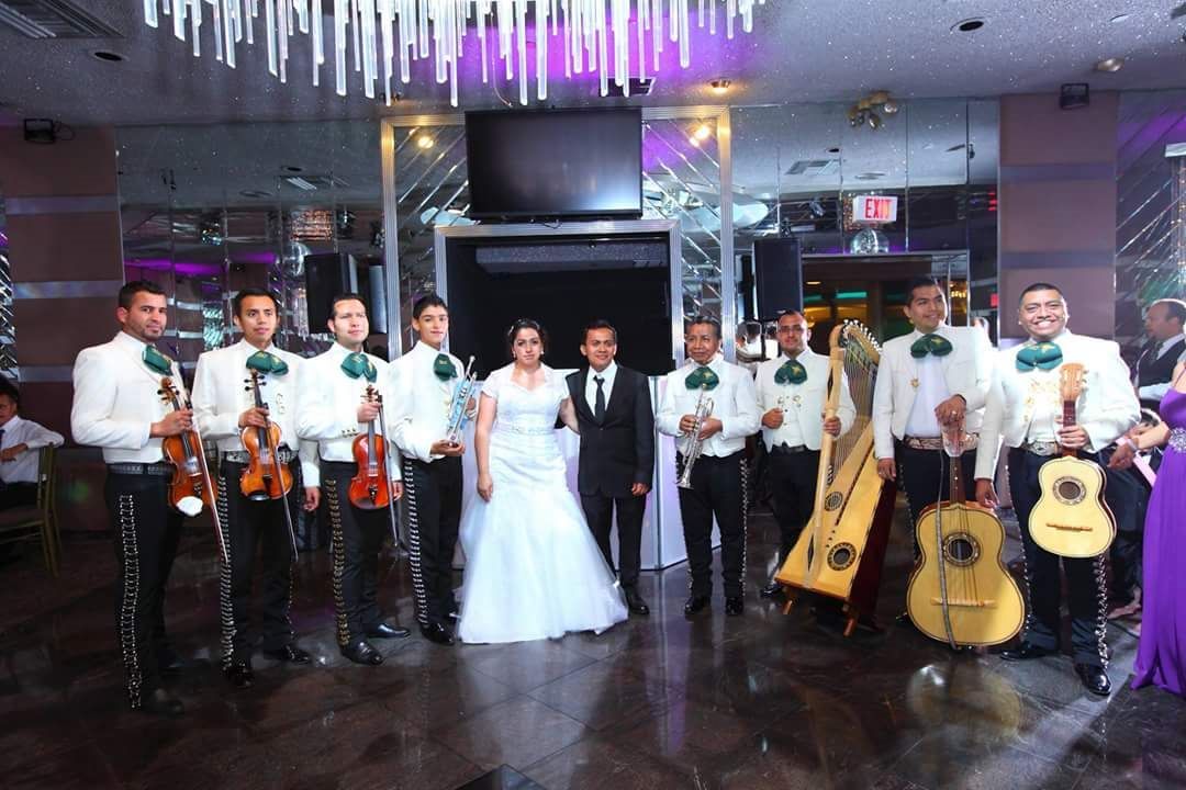 A bride and groom pose in a banquet hall with an eight-member mariachi band playing instruments.