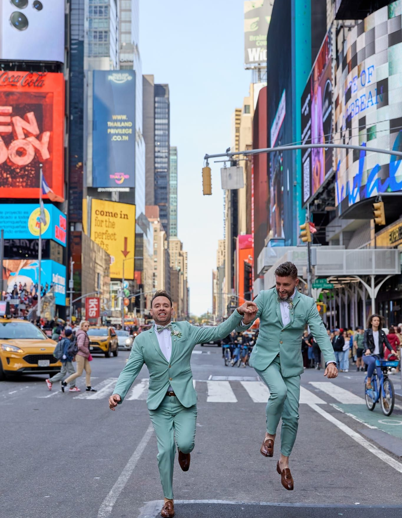 Two people in matching light green suits jump mid-air, holding hands in the middle of a bustling, billboard-lined street.