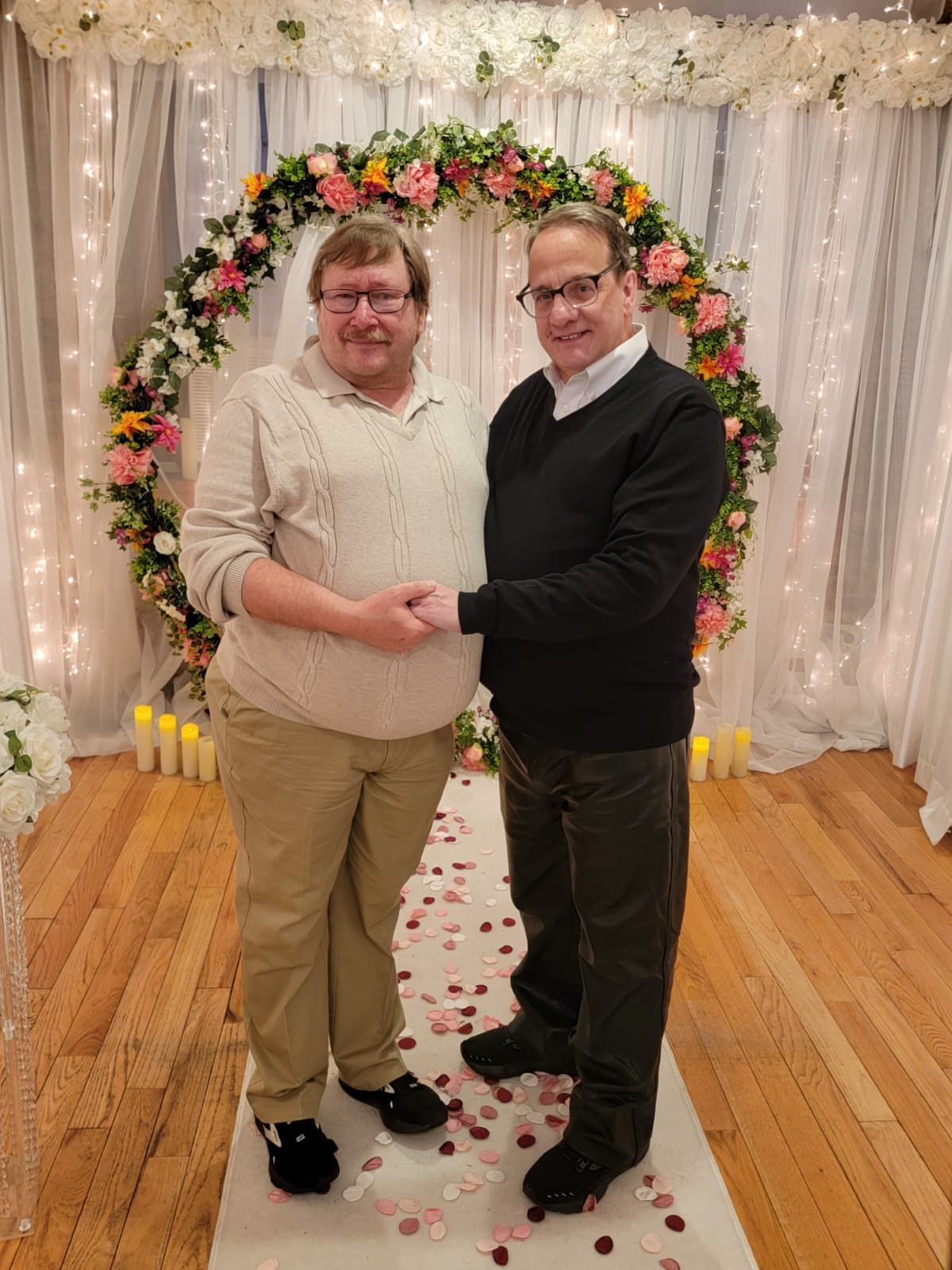 Two people in sweaters stand holding hands in front of a circular floral wedding arch on a decorated floor.