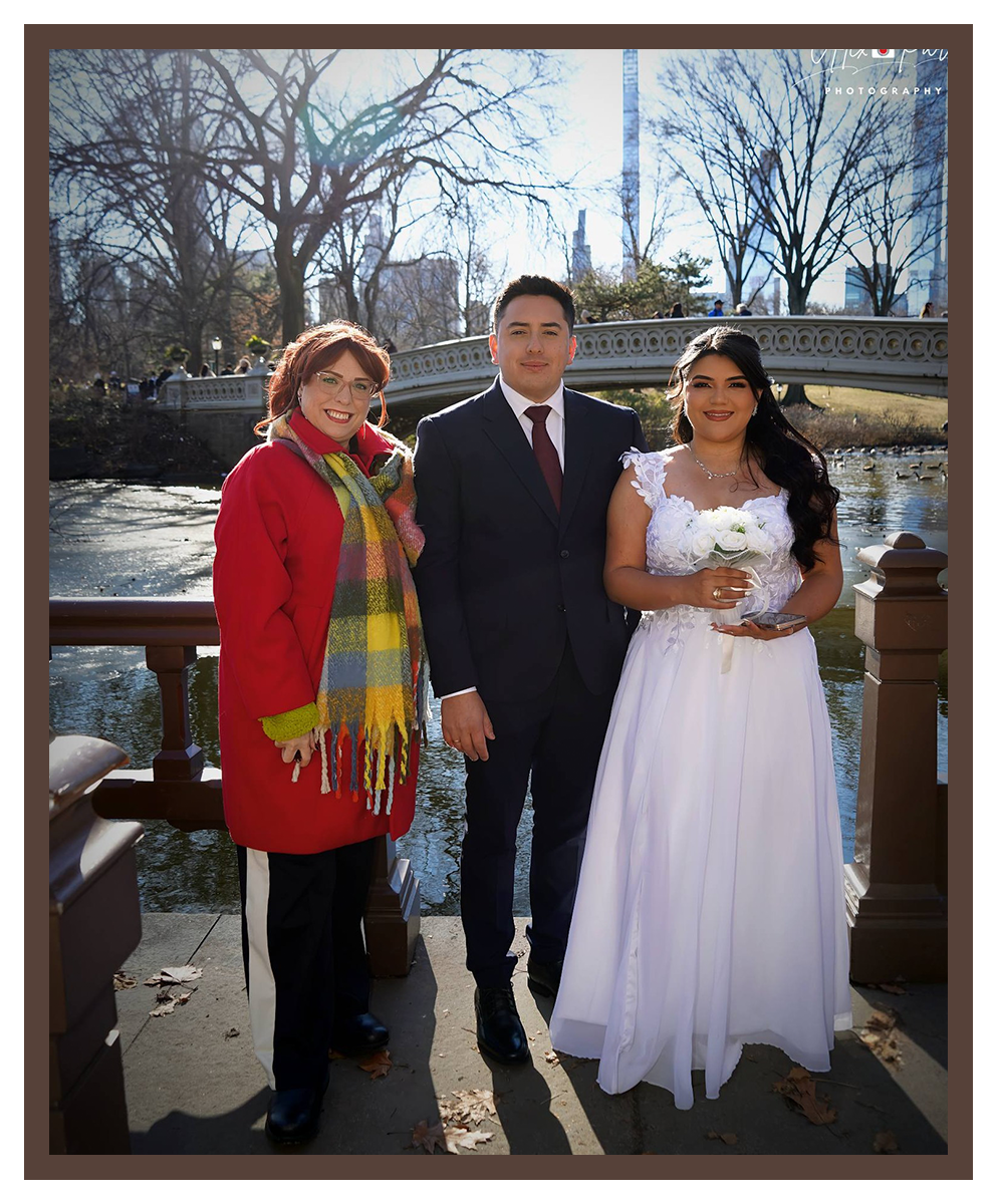 A wedding couple poses for a photo on a park bridge with an officiant, all smiling, with trees and water in the background.