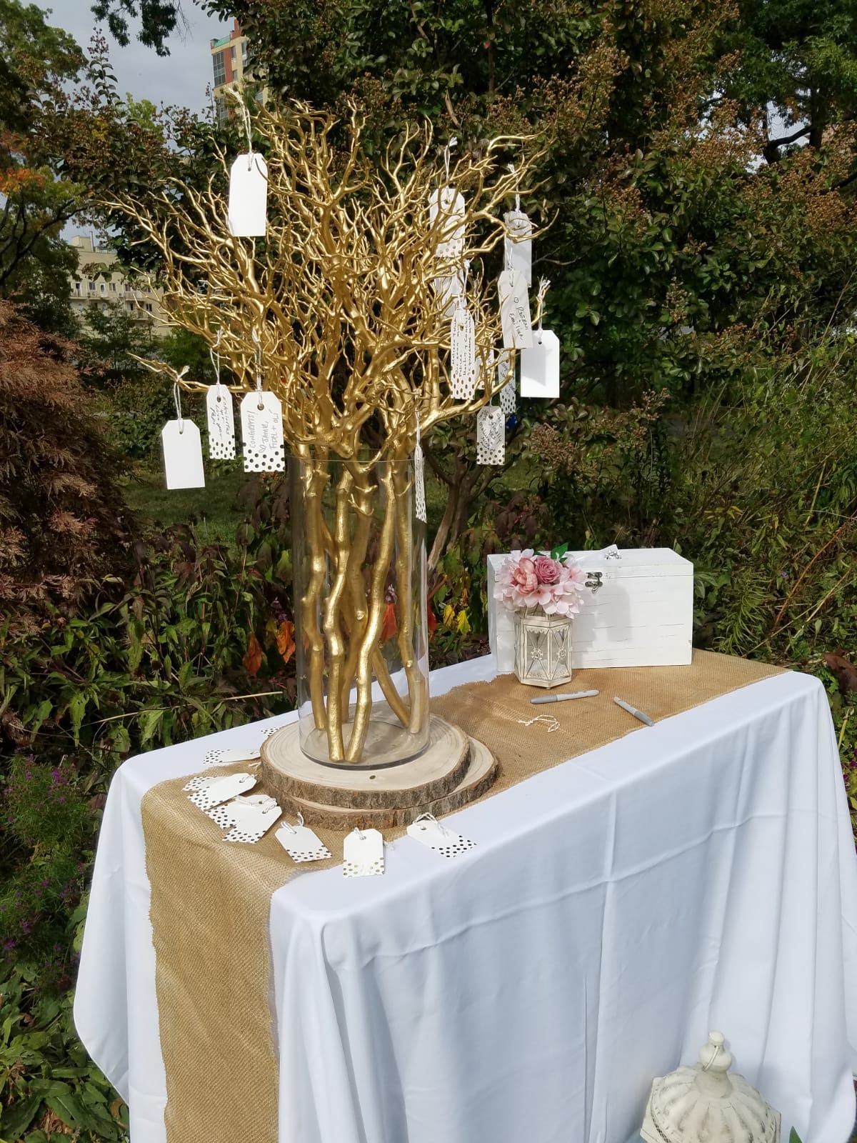 A gold wishing tree with hanging tags on a table covered by a white cloth and burlap runner in a garden setting.