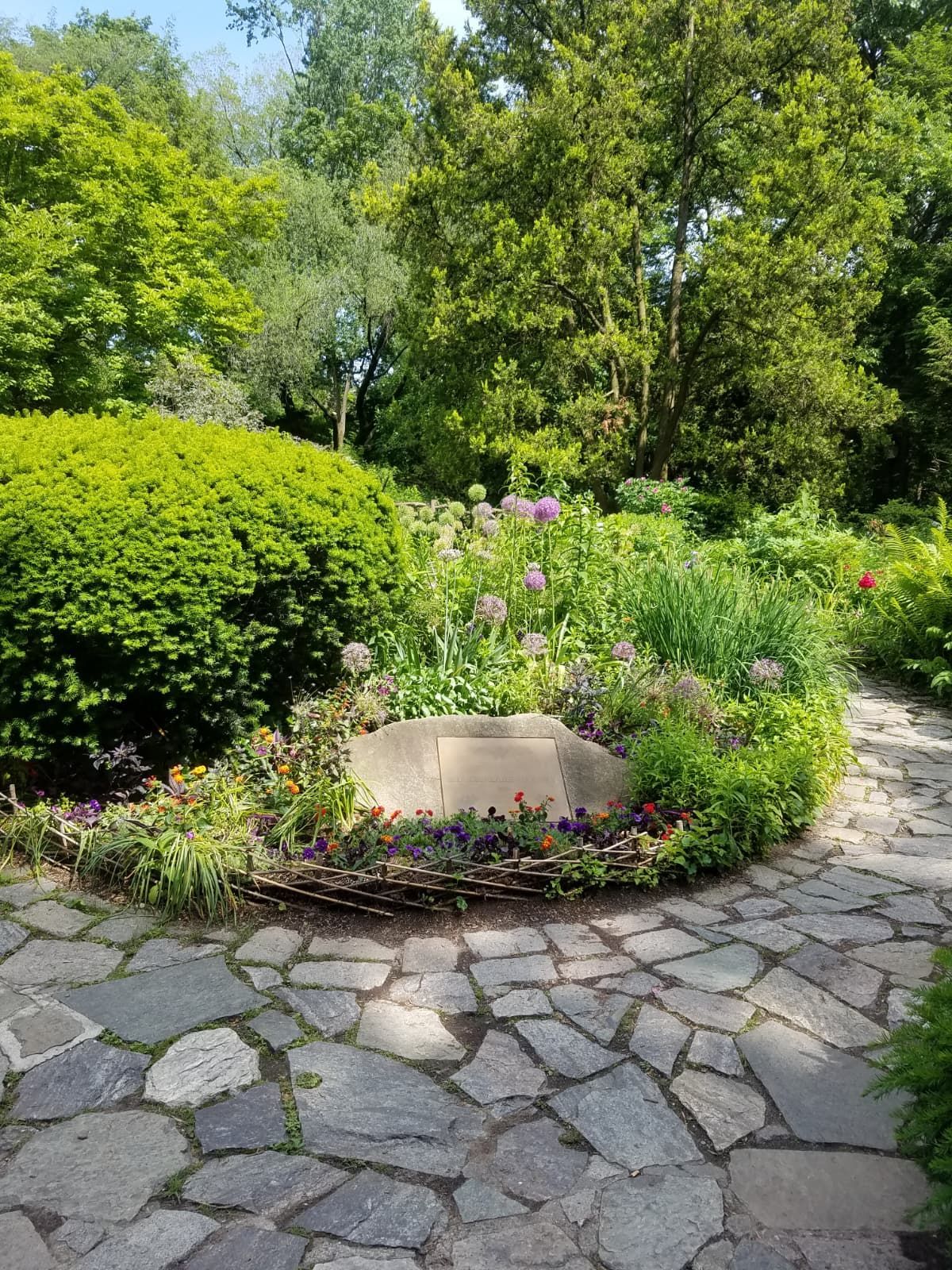 A stone-paved path winds through a lush, sunlit garden featuring a stone monument nestled in a vibrant flower bed.