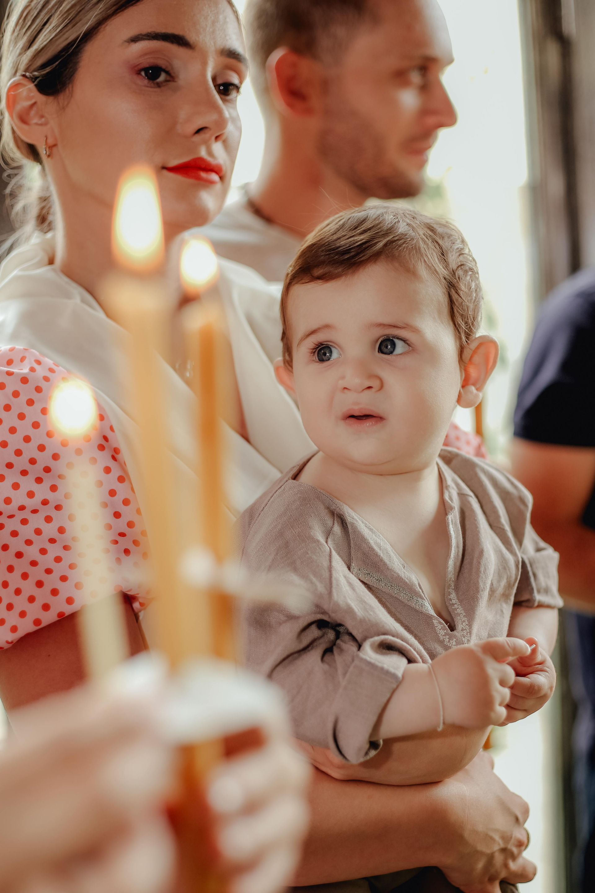 A person holds a toddler during a candlelit ceremony, while another person stands in the background.