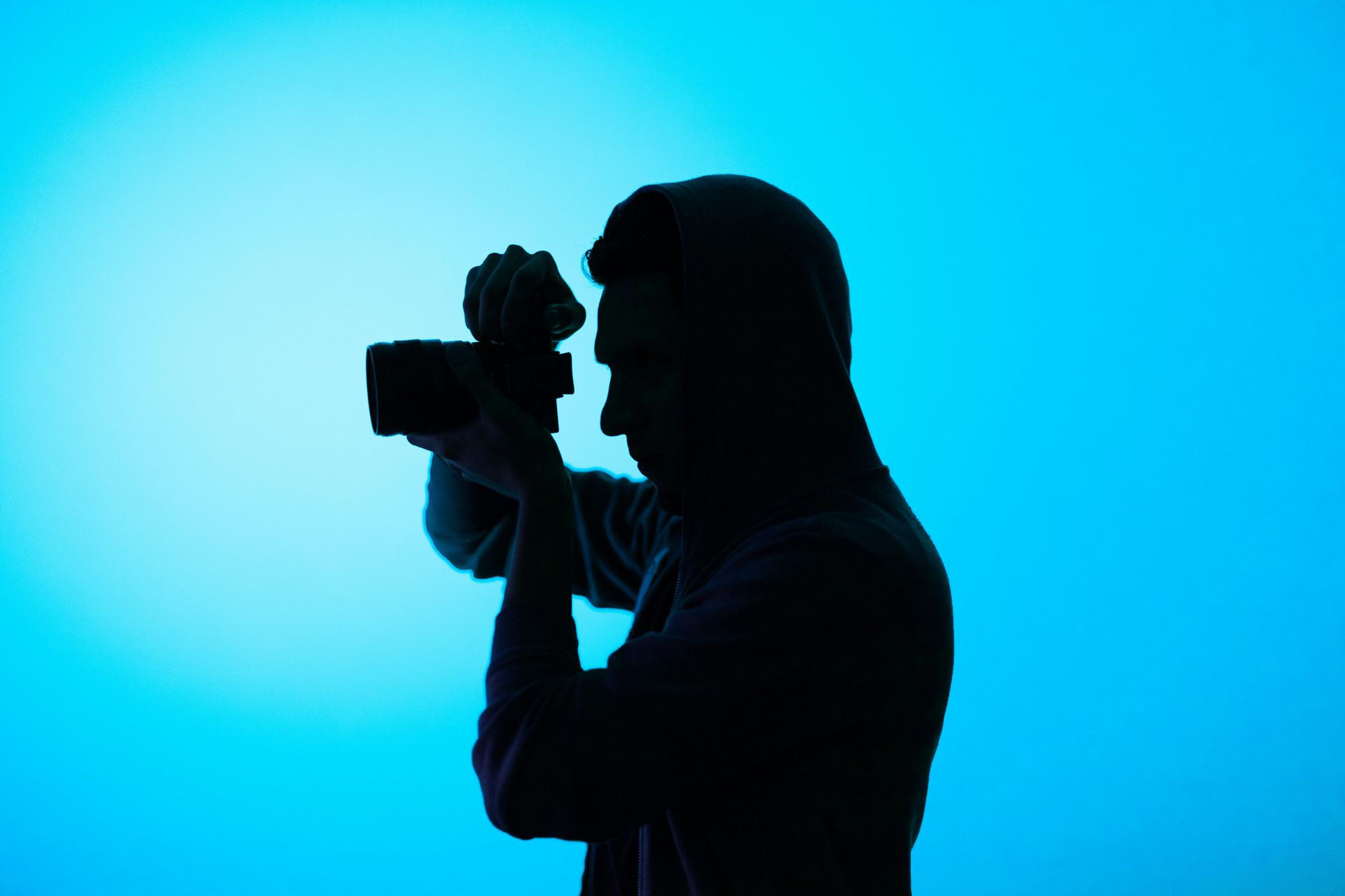 A silhouette of a person wearing a hooded sweatshirt, holding a camera up to their eye against a bright blue background.