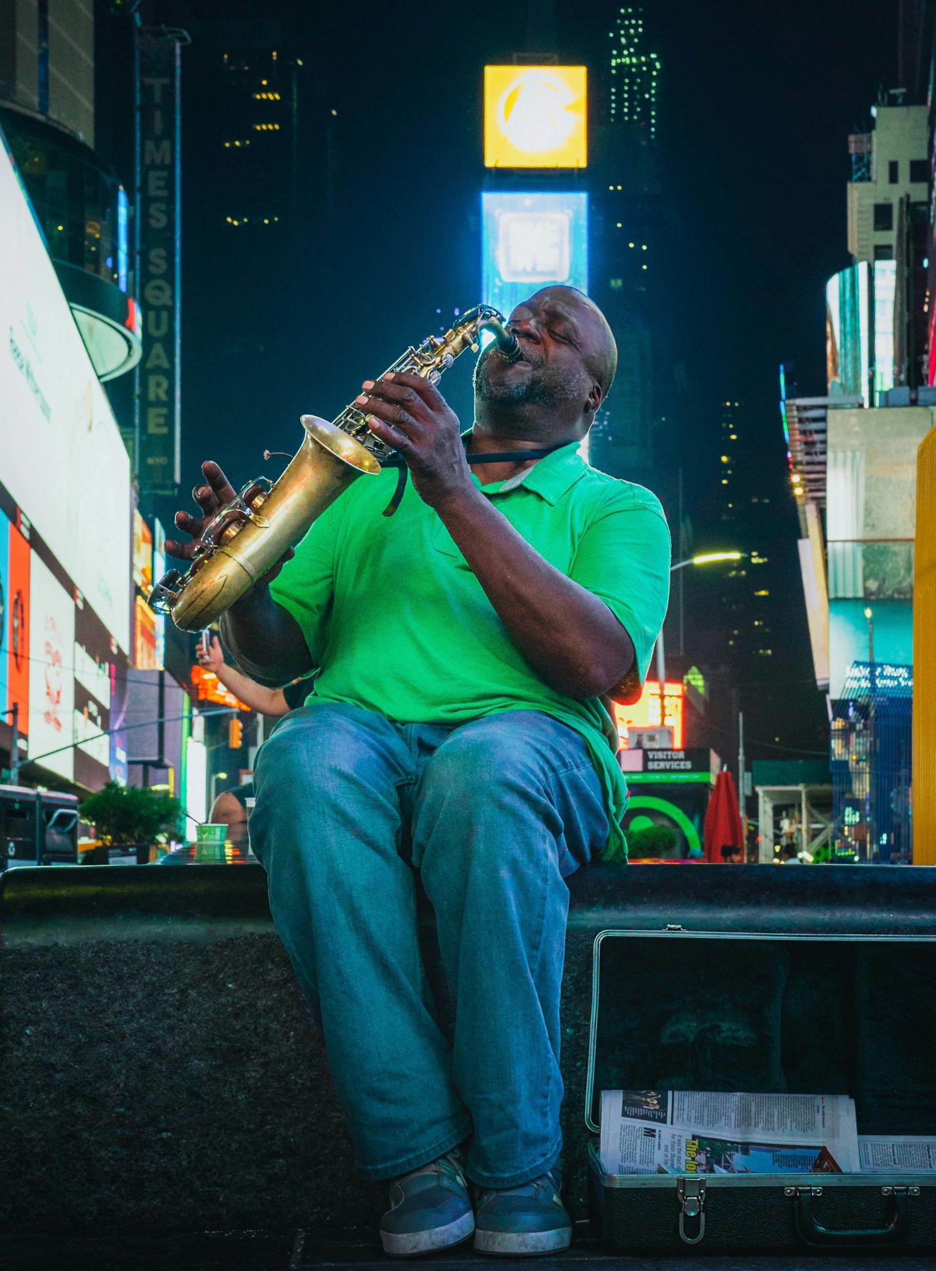 A street musician in a green shirt plays a saxophone while sitting in Times Square at night.
