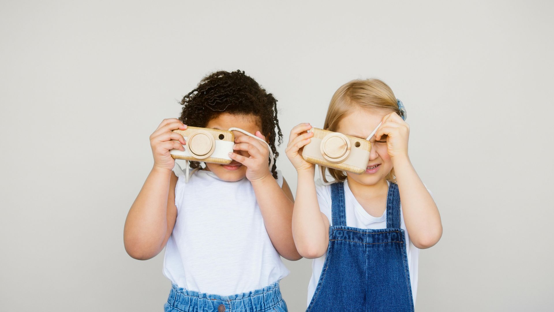 Two people standing side-by-side, each holding a wooden toy camera up to their face against a light grey background.
