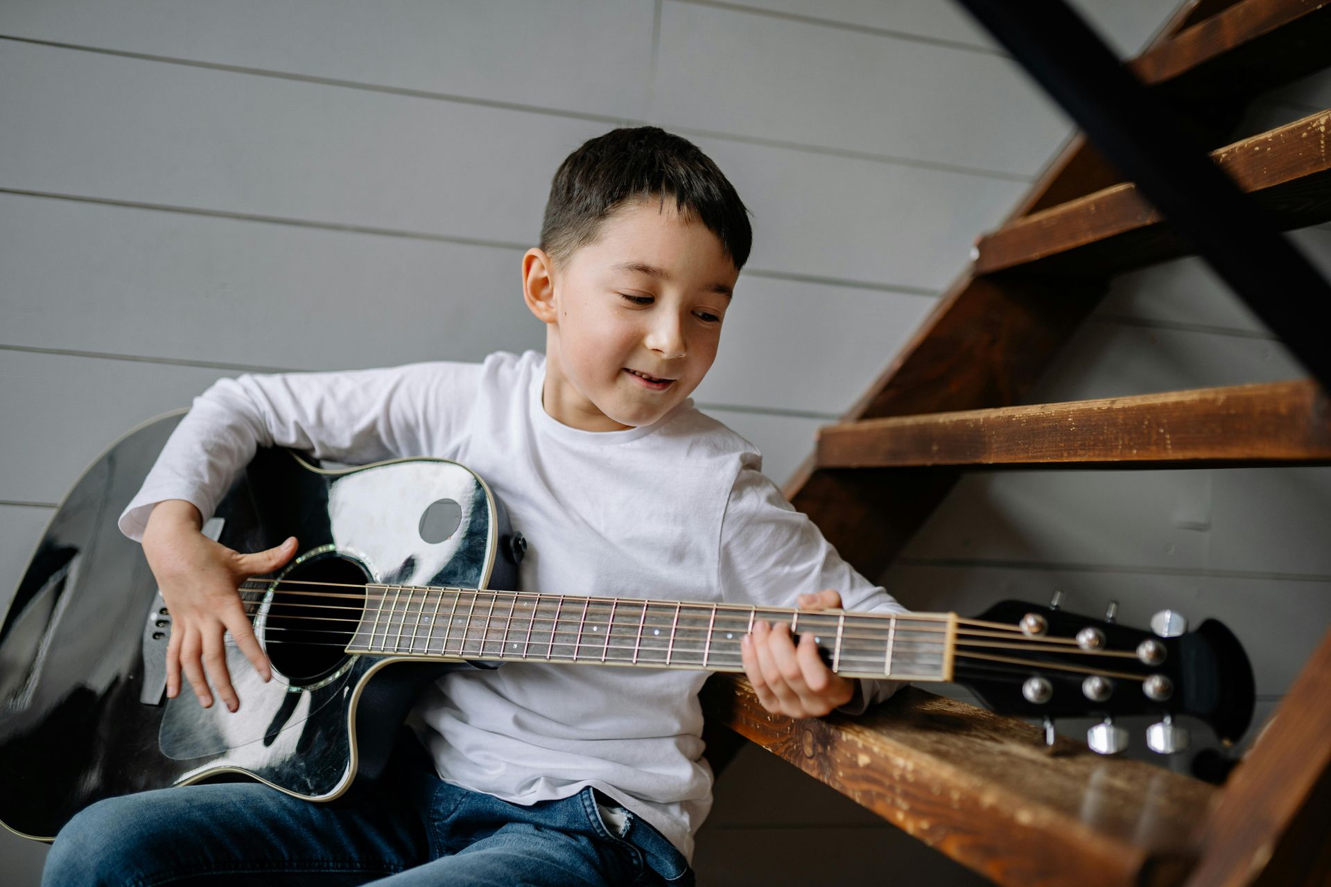 A young person in a white long-sleeved shirt plays an acoustic guitar while sitting on wooden stairs.