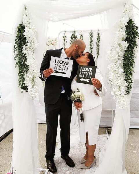 A couple kisses under a white wedding arch, holding signs that say 