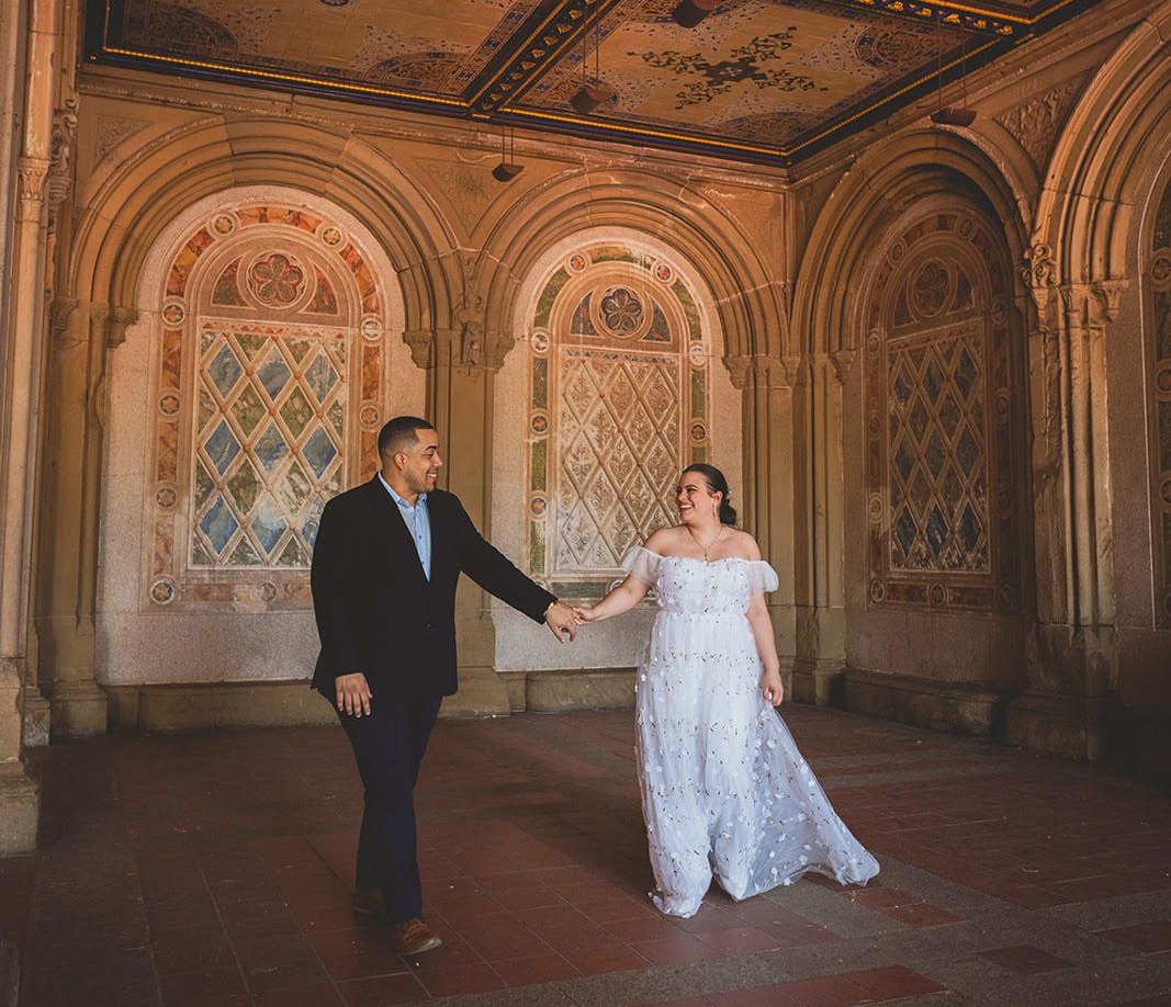 A couple in formal attire walks hand-in-hand through the ornate, tiled arcade of Bethesda Terrace in Central Park.